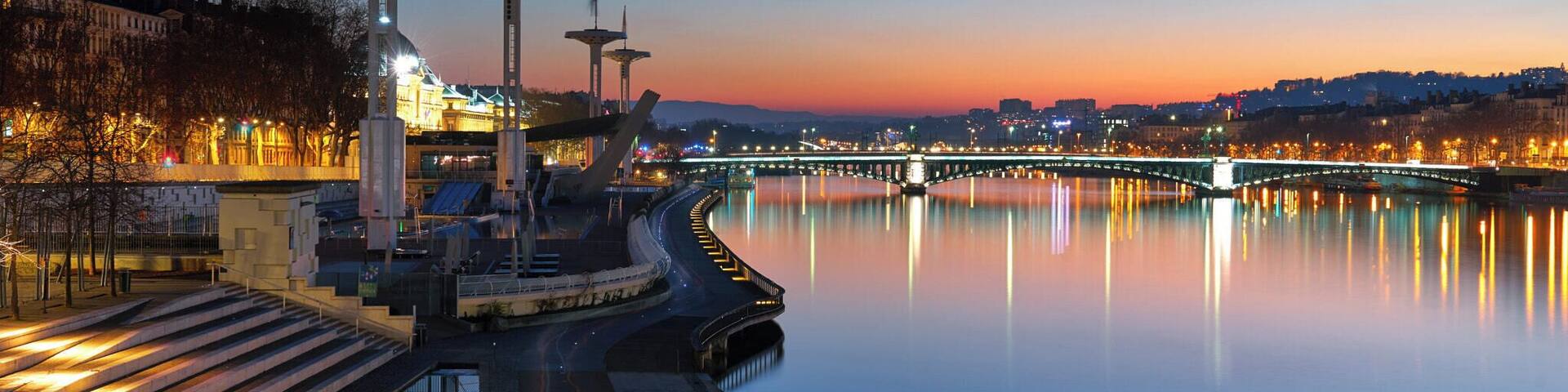 A calm river with a bridge, illuminated structures, and a cityscape in the background.