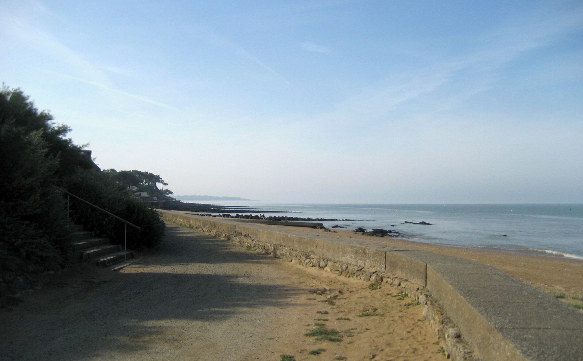 A coastal road with a sandy beach, a stone wall, and a clear sky.