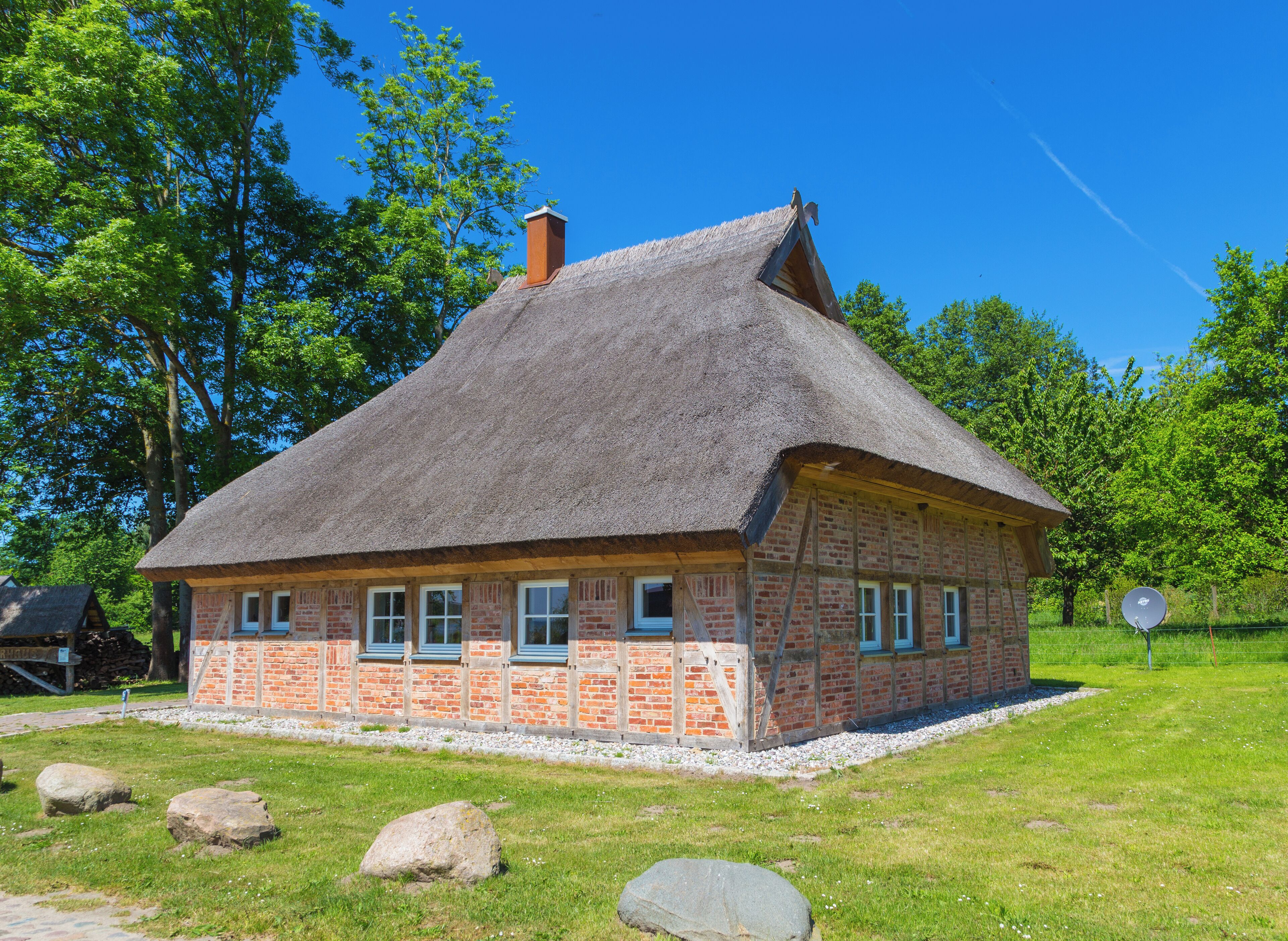 A traditional thatched-roof house with brick walls, surrounded by a grassy area with rocks and trees.