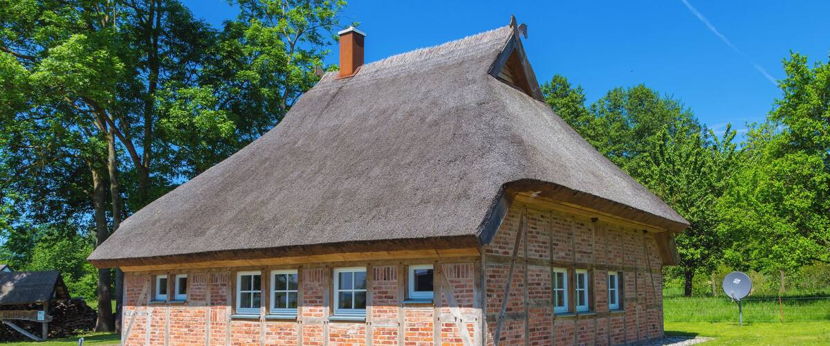 A traditional thatched-roof house with brick walls, surrounded by a grassy area with rocks and trees.