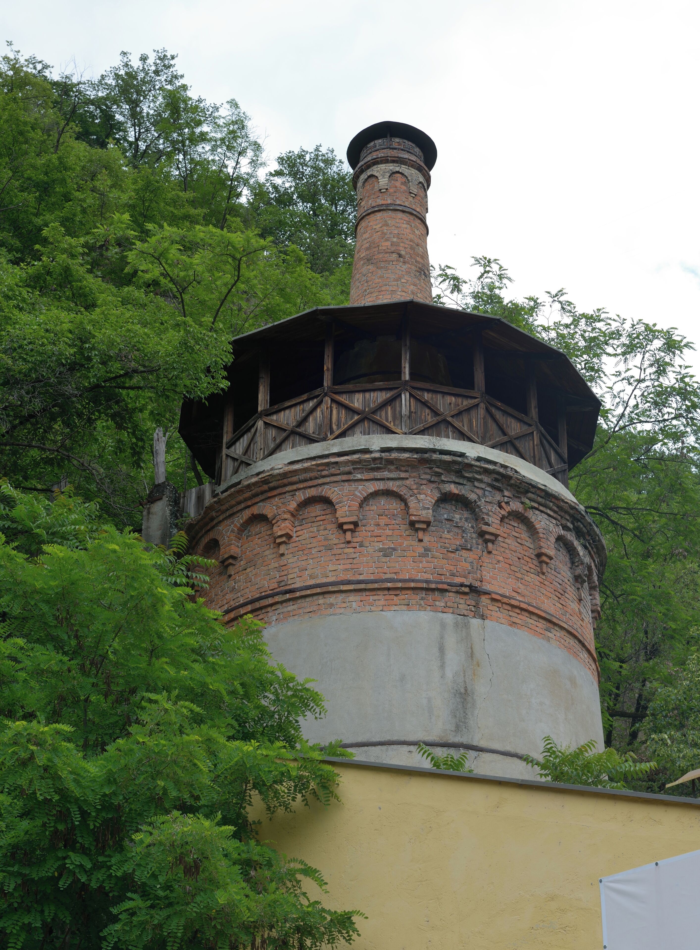 A brick tower with a wooden balcony, surrounded by lush green trees.