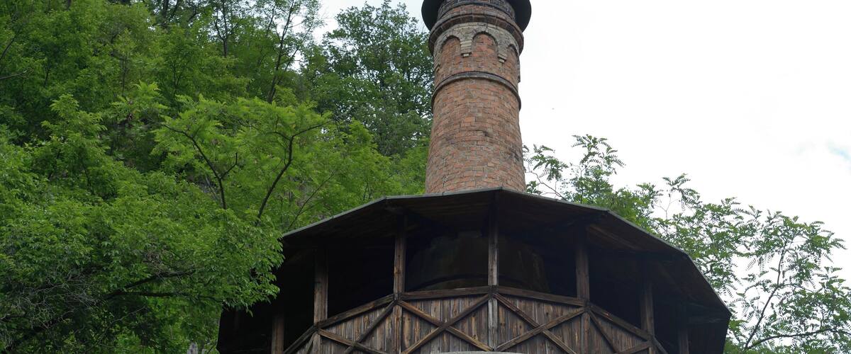 A brick tower with a wooden balcony, surrounded by lush green trees.