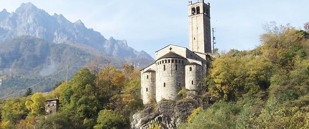 A stone church with a bell tower on a hillside, surrounded by autumn foliage and a river.