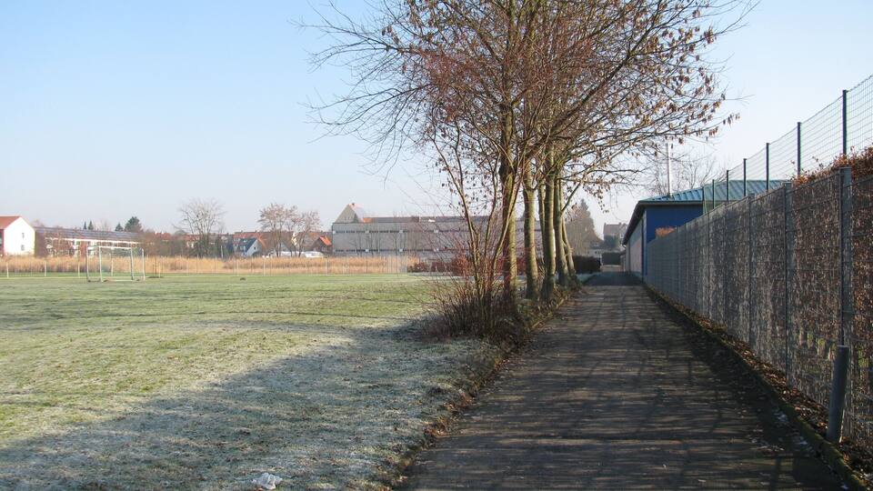 A pathway alongside a sports field with a fence and leafless trees.