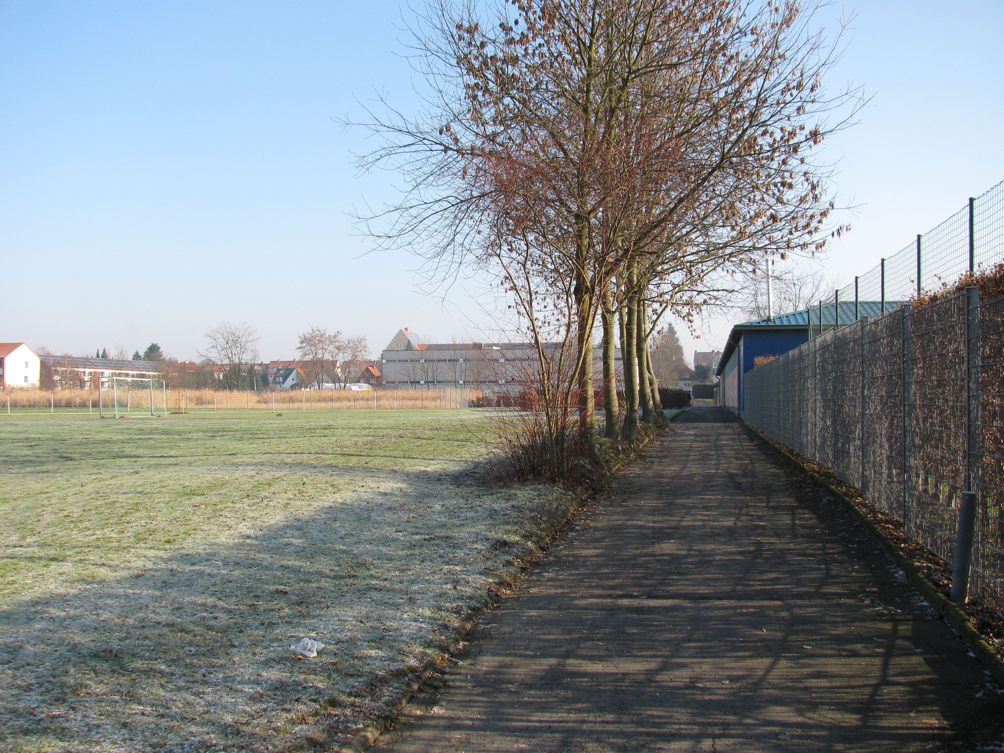 A pathway alongside a sports field with a fence and leafless trees.