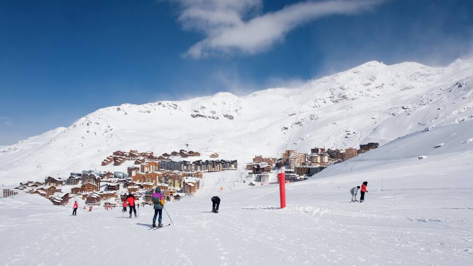 A snowy ski slope with skiers, a red pole, and a town in the background.
