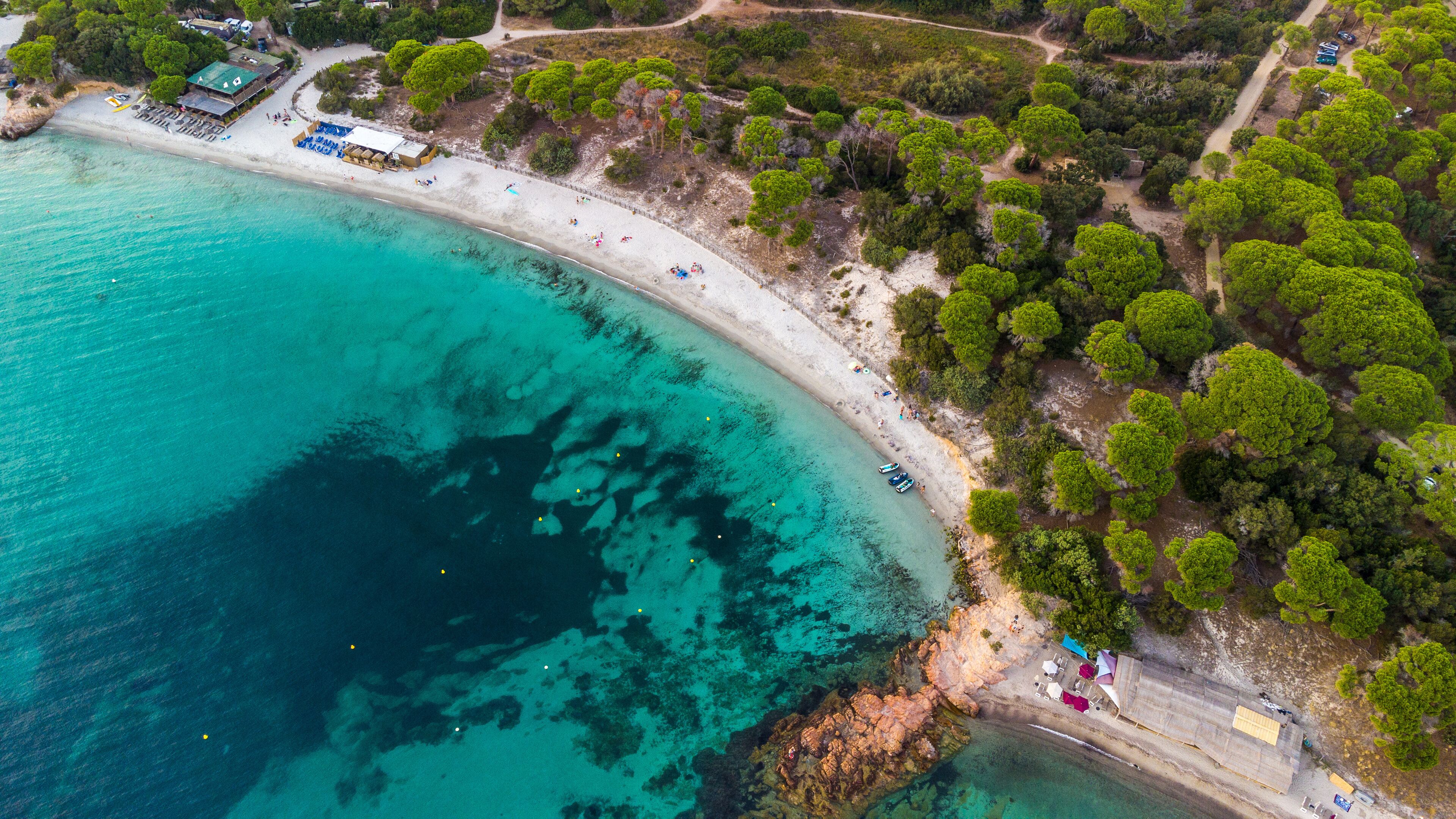 An aerial view of a coastal area with a sandy beach, clear turquoise waters, and dense green trees.