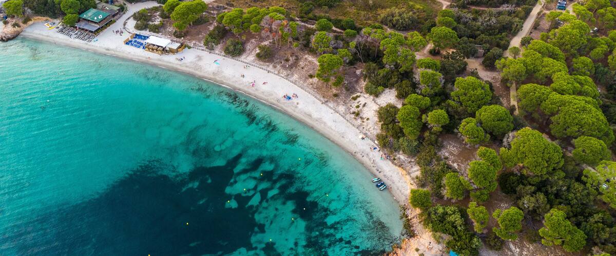 An aerial view of a coastal area with a sandy beach, clear turquoise waters, and dense green trees.