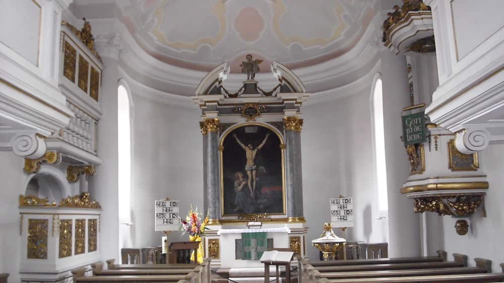 Ein aufwendig gestaltetes Kircheninnere mit Kruzifix, Kirchenbänken und dekorativen Säulen.
