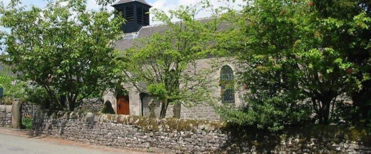 A stone church with a bell tower, surrounded by trees and a stone wall.