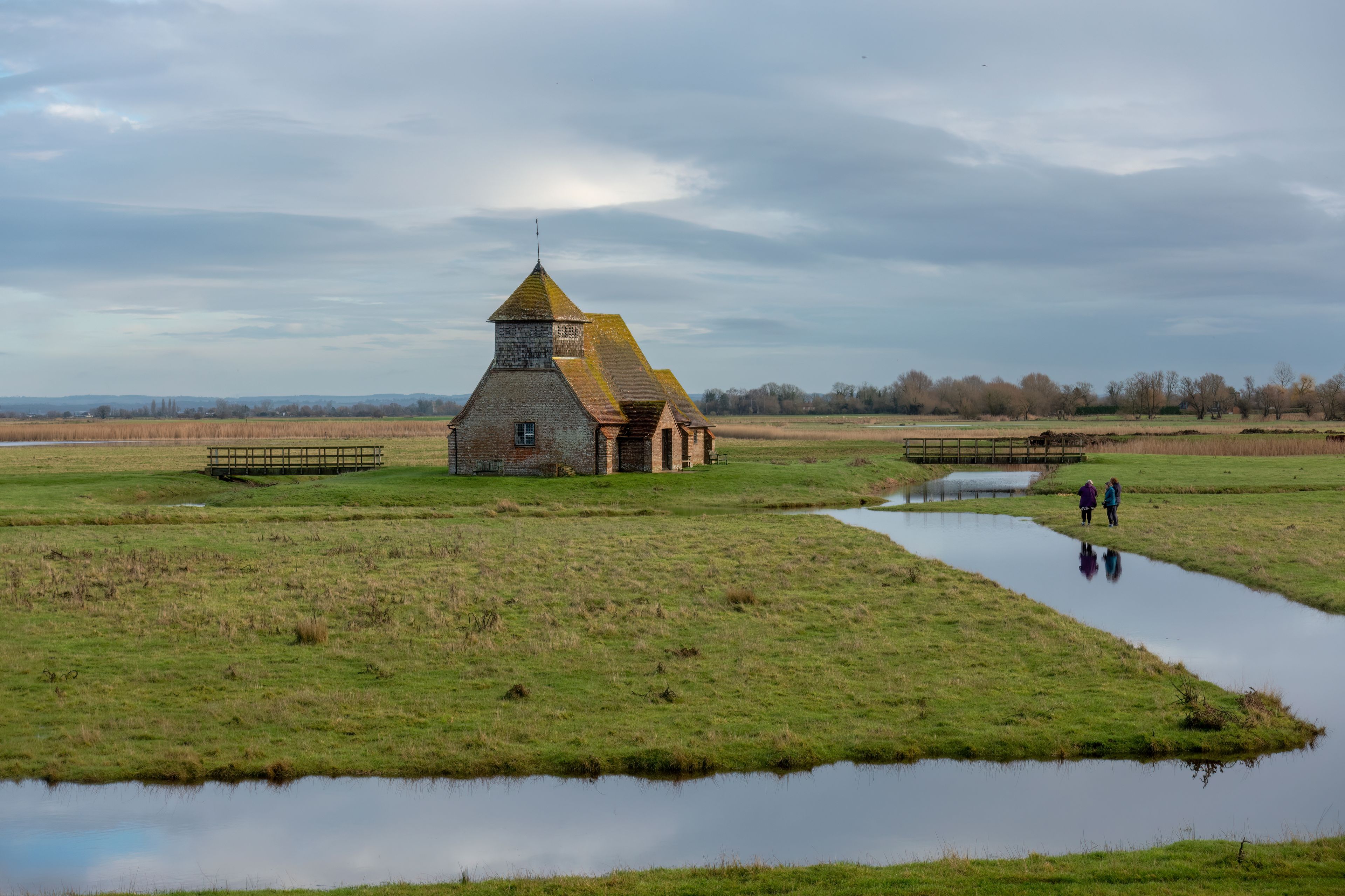A rural landscape with a traditional brick building featuring a conical roof, surrounded by open fields and a small water channel.