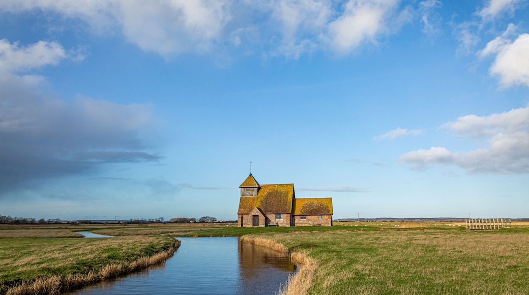 A rural landscape with a small church, a narrow water channel, and a clear blue sky.