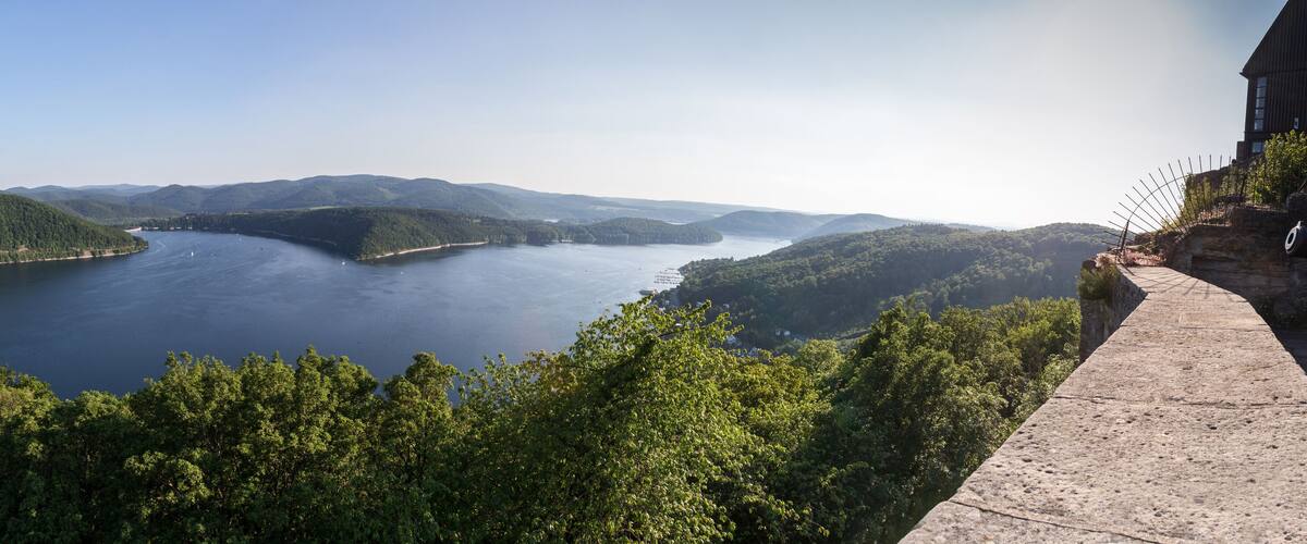 A panoramic view of a lake surrounded by forested hills and a stone bridge.