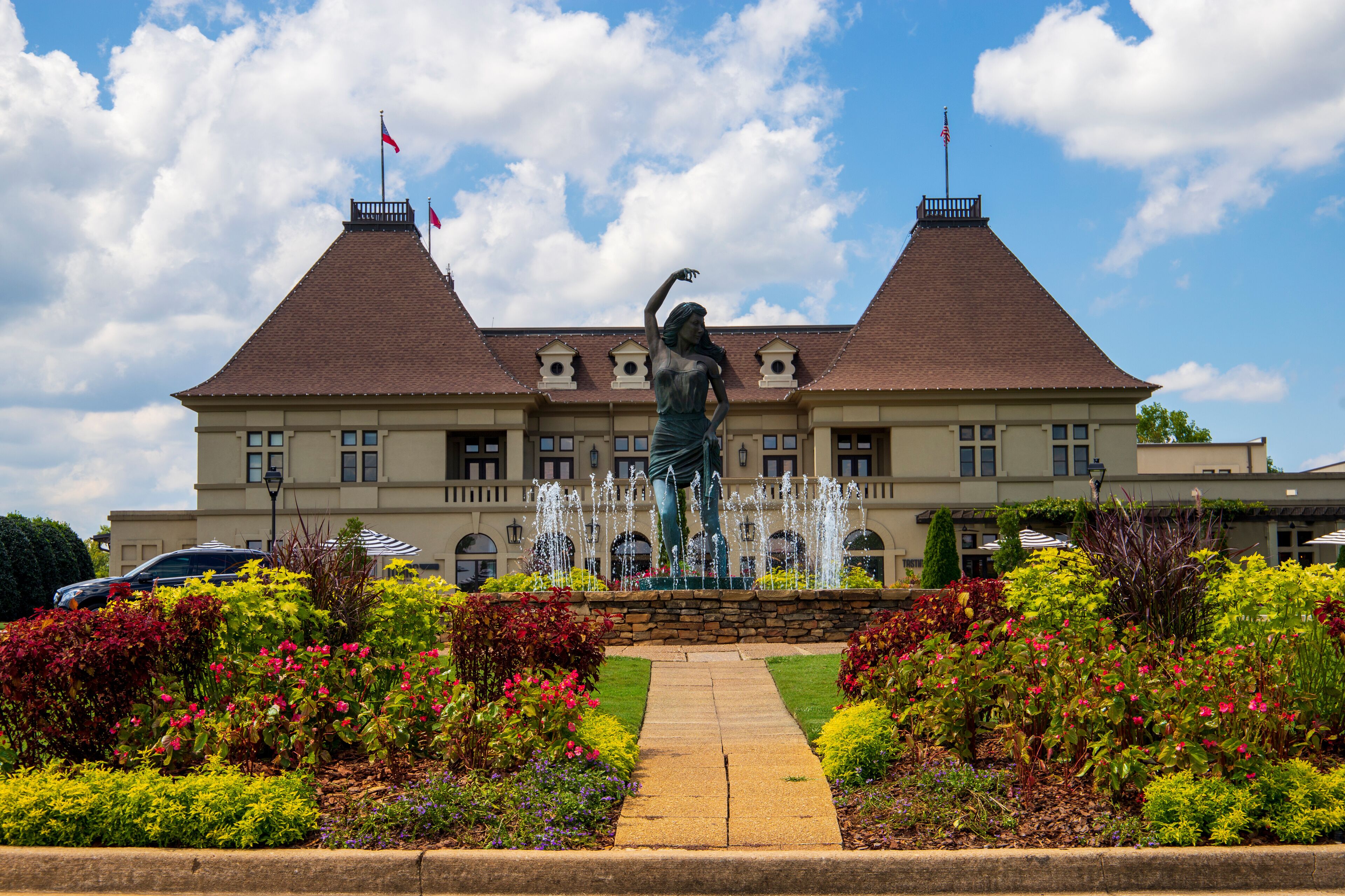 a gorgeous summer landscape with a water fountain with statue of a woman in the center surrounded by colorful flowers and lush green trees, grass and plants with blue sky and powerful clouds