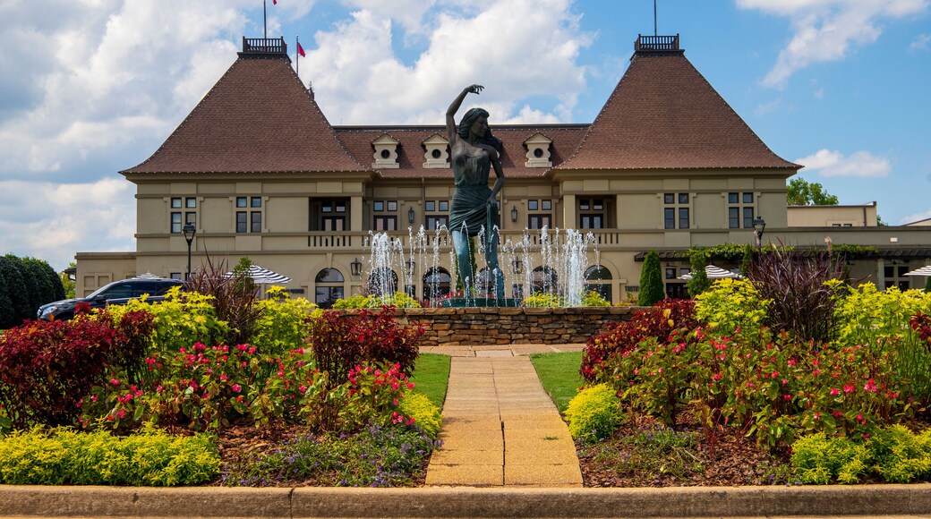 a gorgeous summer landscape with a water fountain with statue of a woman in the center surrounded by colorful flowers and lush green trees, grass and plants with blue sky and powerful clouds