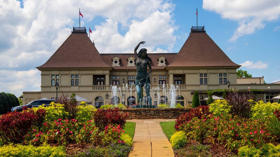 a gorgeous summer landscape with a water fountain with statue of a woman in the center surrounded by colorful flowers and lush green trees, grass and plants with blue sky and powerful clouds