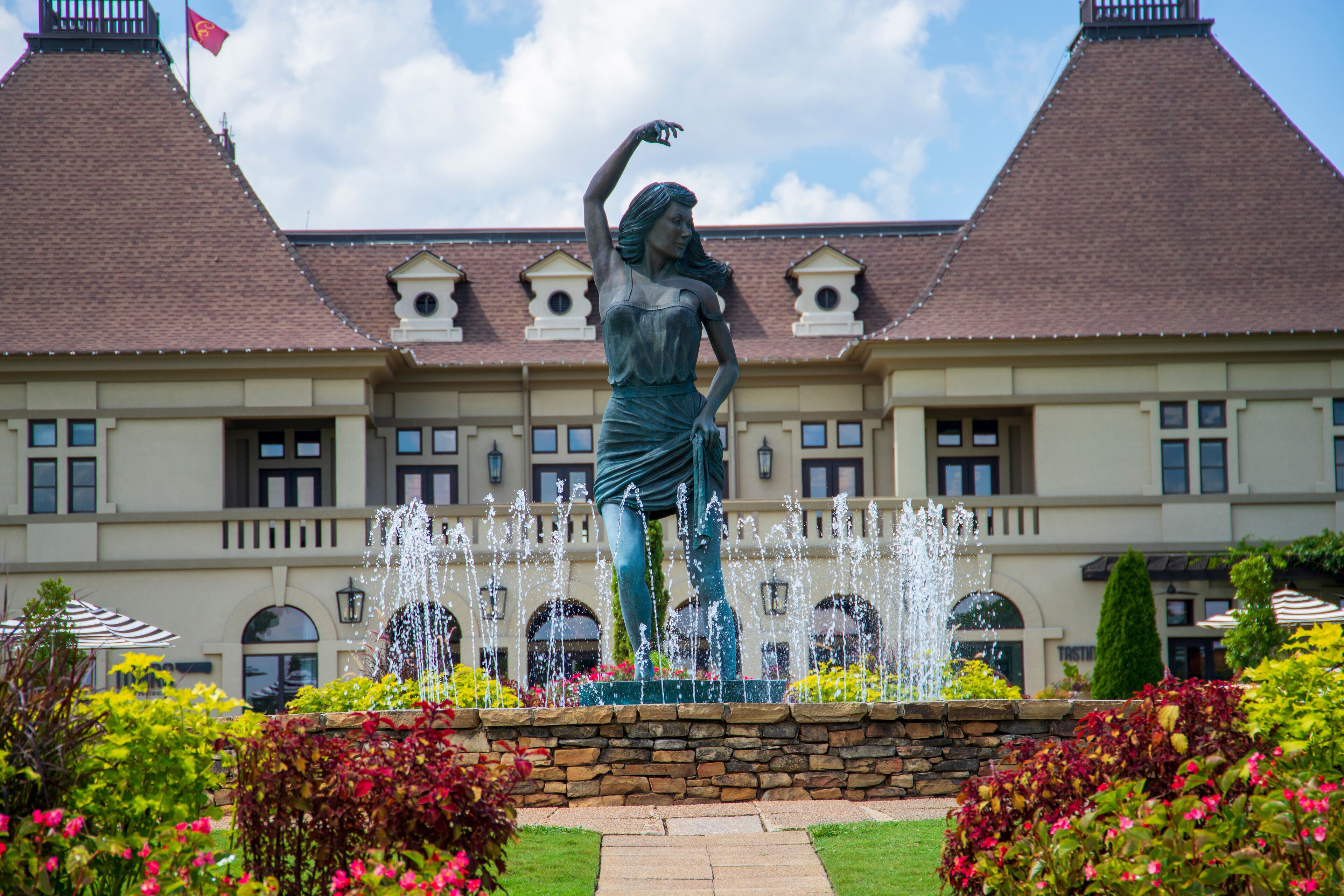a gorgeous summer landscape with a water fountain with statue of a woman in the center surrounded by colorful flowers and lush green trees, grass and plants with blue sky and powerful clouds