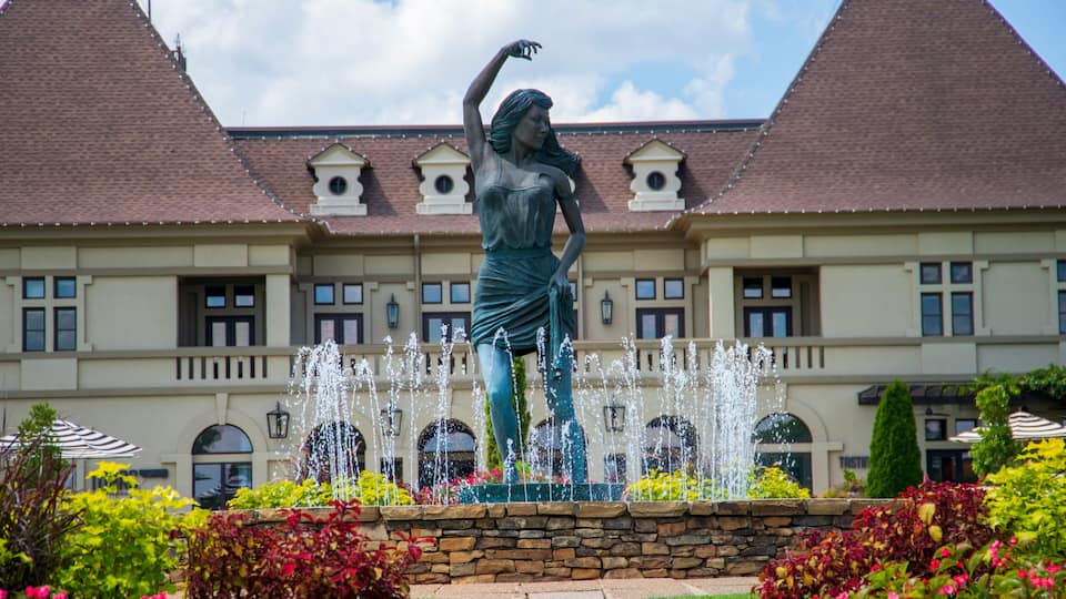 a gorgeous summer landscape with a water fountain with statue of a woman in the center surrounded by colorful flowers and lush green trees, grass and plants with blue sky and powerful clouds