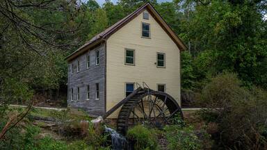 A historic water mill with a wooden structure and a large water wheel, surrounded by dense greenery.