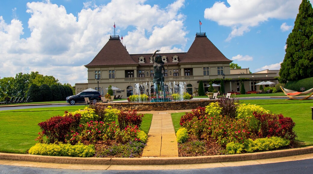 a gorgeous summer landscape with a water fountain with statue of a woman in the center surrounded by colorful flowers and lush green trees, grass and plants with blue sky and powerful clouds