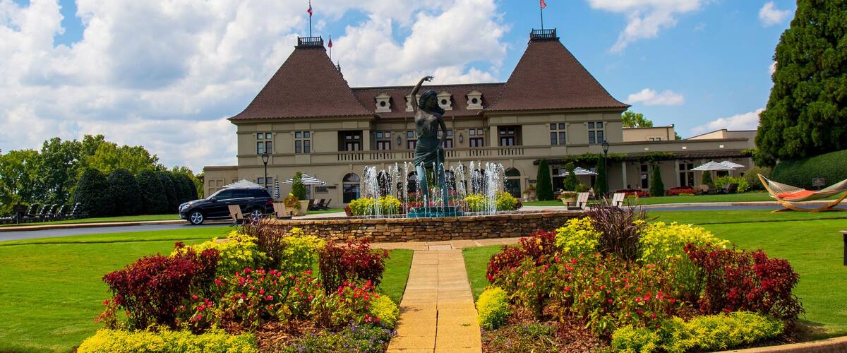 a gorgeous summer landscape with a water fountain with statue of a woman in the center surrounded by colorful flowers and lush green trees, grass and plants with blue sky and powerful clouds