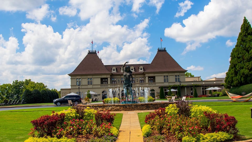 a gorgeous summer landscape with a water fountain with statue of a woman in the center surrounded by colorful flowers and lush green trees, grass and plants with blue sky and powerful clouds