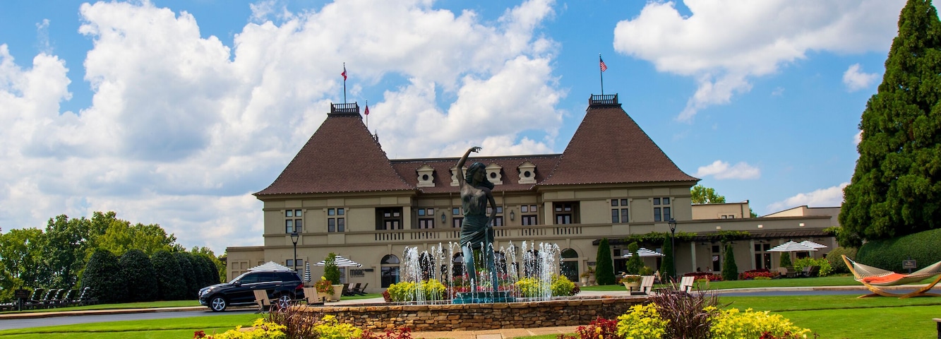 a gorgeous summer landscape with a water fountain with statue of a woman in the center surrounded by colorful flowers and lush green trees, grass and plants with blue sky and powerful clouds