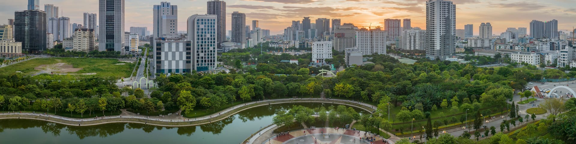 A cityscape with a river, green park, and high-rise buildings during sunset.