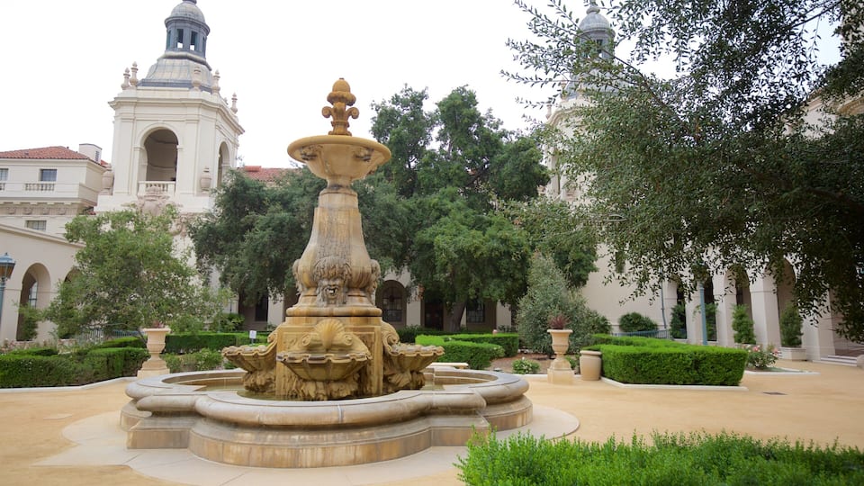 A classical fountain with ornate lion sculptures in a courtyard surrounded by manicured hedges and trees, with a white building featuring a dome in the background.