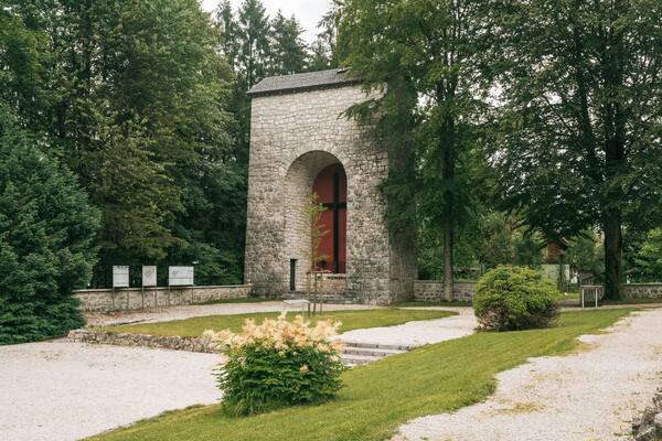 A stone memorial with an arched entrance, surrounded by lush greenery and a gravel path.