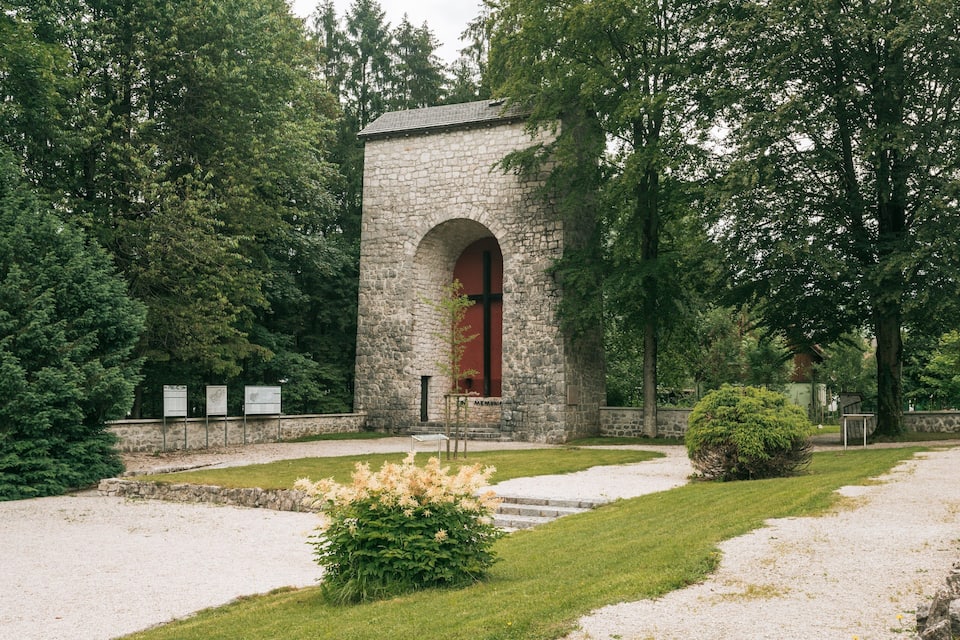 A stone memorial with an arched entrance, surrounded by lush greenery and a gravel path.