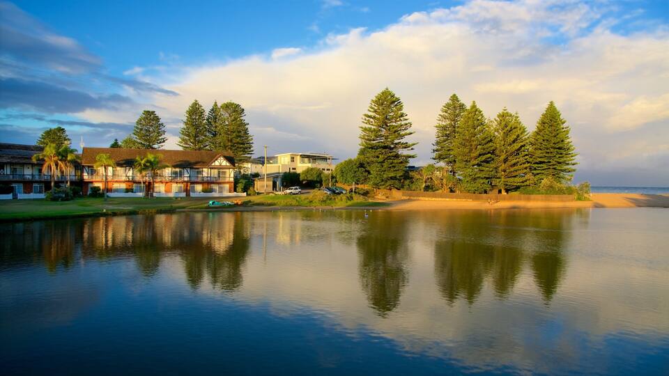 A calm lake with reflections of trees and buildings, a sandy beach, and a clear sky.