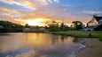 A sunset over a calm river with a bridge, a lodge, and surrounding greenery.