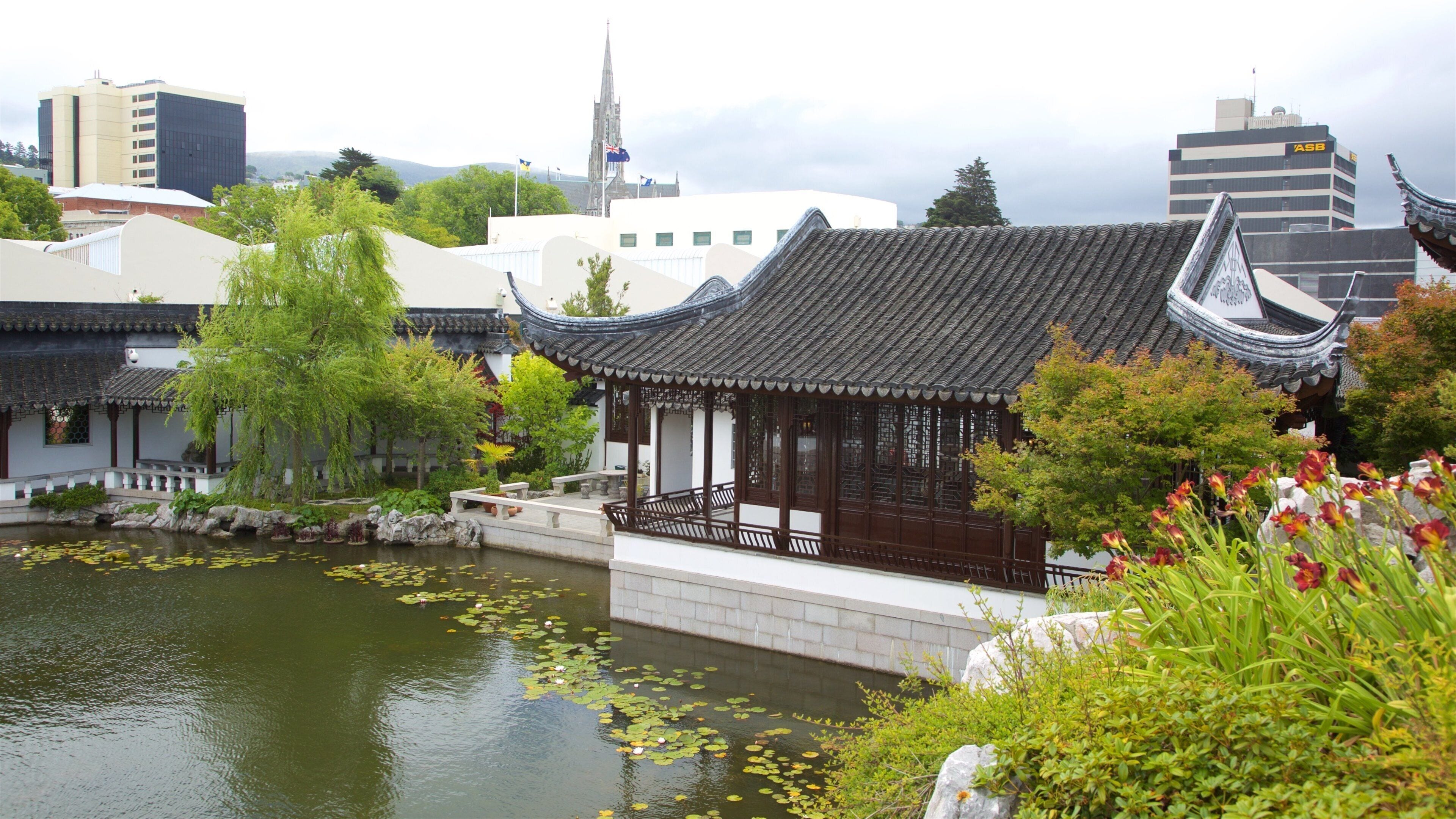 Un pavillon chinois traditionnel, situé au bord d’un étang parsemé de nénuphars, entouré de verdure et avec des bâtiments modernes en arrière-plan.