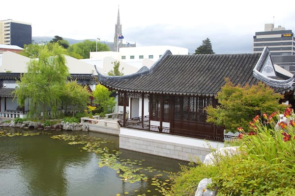 Um pavilhão chinês tradicional à beira de um lago com folhas de nenúfar, rodeado por vegetação e edifícios modernos ao fundo.