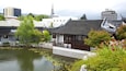 A traditional Chinese pavilion by a pond with lily pads, surrounded by greenery and modern buildings in the background.