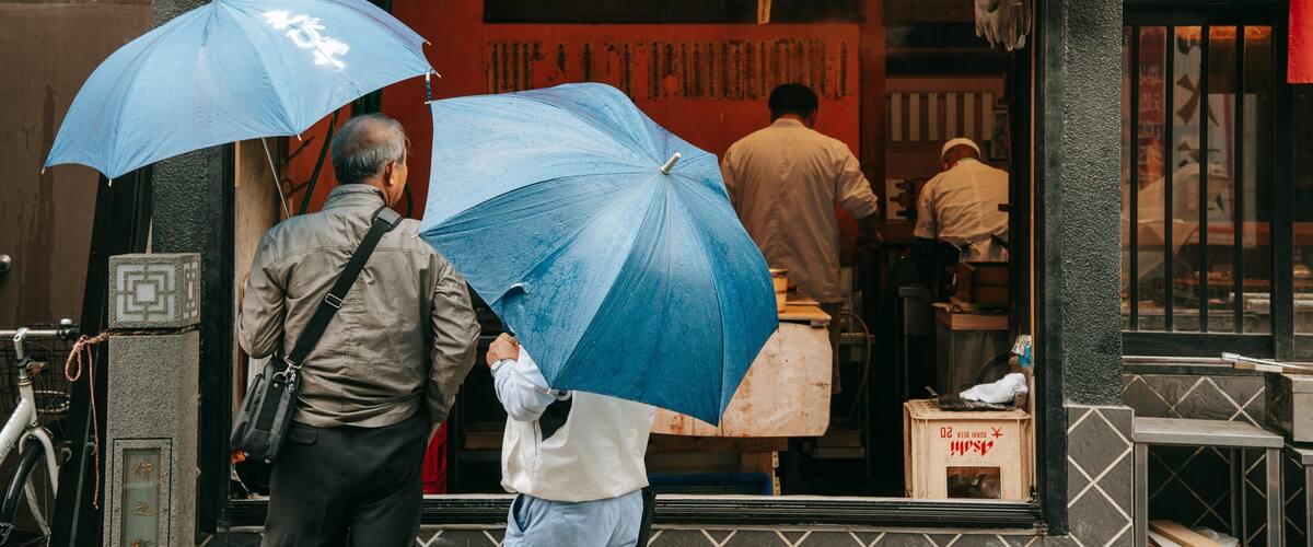 Two people holding blue umbrellas at a restaurant entrance with a red awning and Japanese characters.
