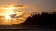 A sunset over a coastal landscape with a forested hill and ocean.