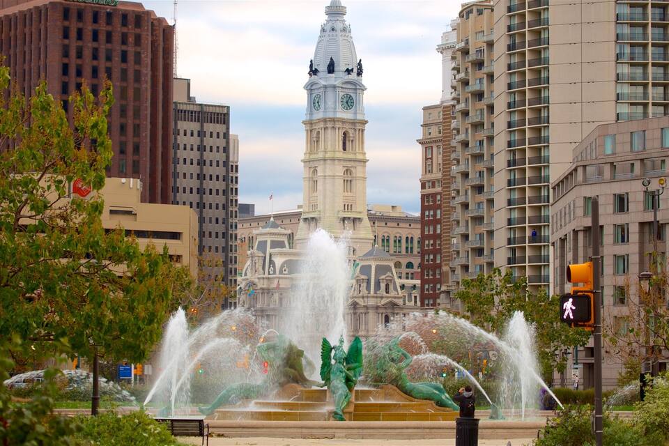 A city fountain with water jets and statues, surrounded by tall buildings and a clock tower.