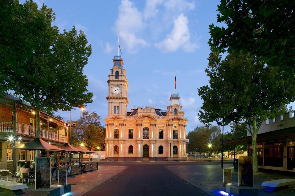 A historic building with two towers and a clock, surrounded by trees and a plaza with outdoor seating.