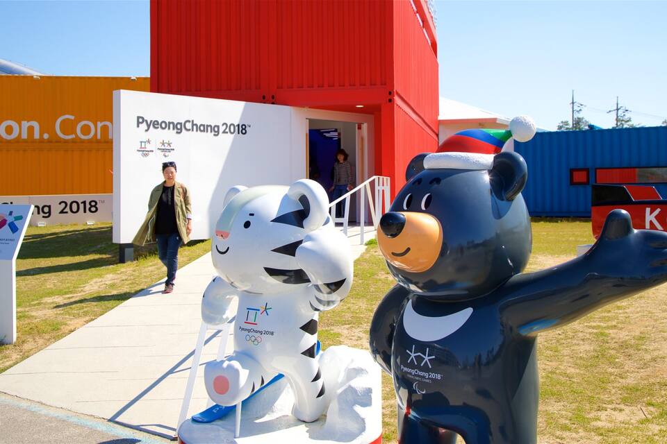Two mascots, a white bear and a blue bear, in front of a building with a sign that reads "PyeongChang 2018".
