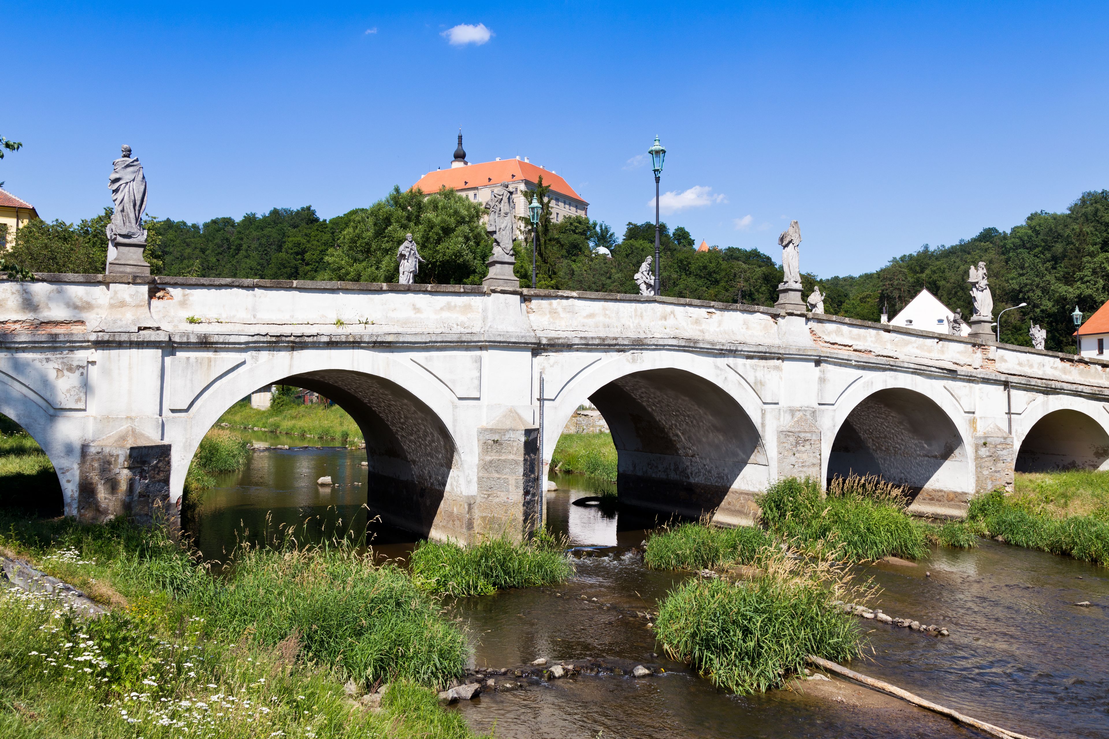 Eine Steinbrücke mit mehreren Bögen über einem Fluss, Statuen auf der Brücke und ein Gebäude mit rotem Dach im Hintergrund.
