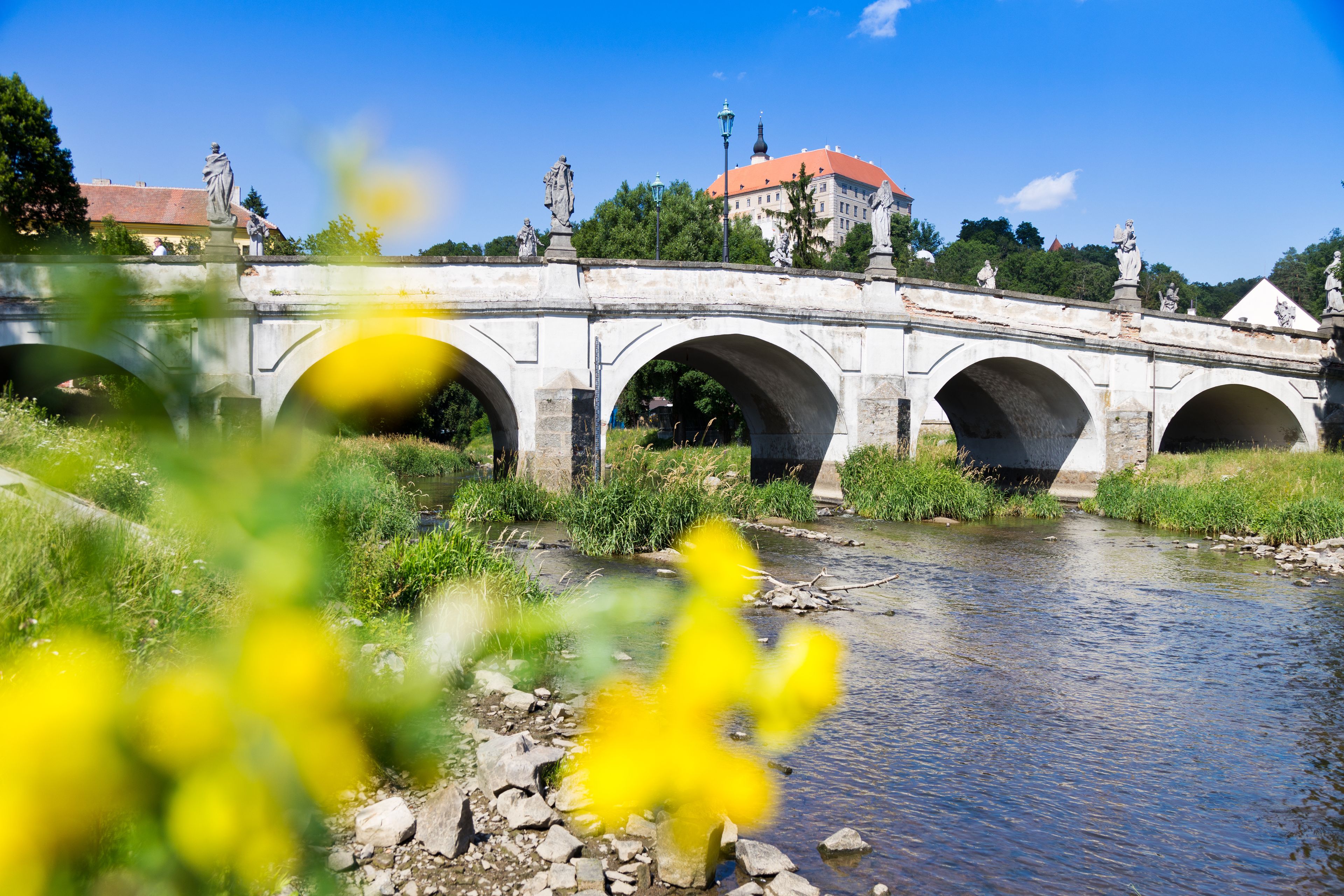 Eine Steinbrücke mit mehreren Bögen über einem Fluss, umgeben von Grünpflanzen und einem Gebäude mit rotem Dach im Hintergrund.