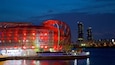 A modern red-lit building with a unique curved roof, situated by a waterfront with a city skyline in the background.