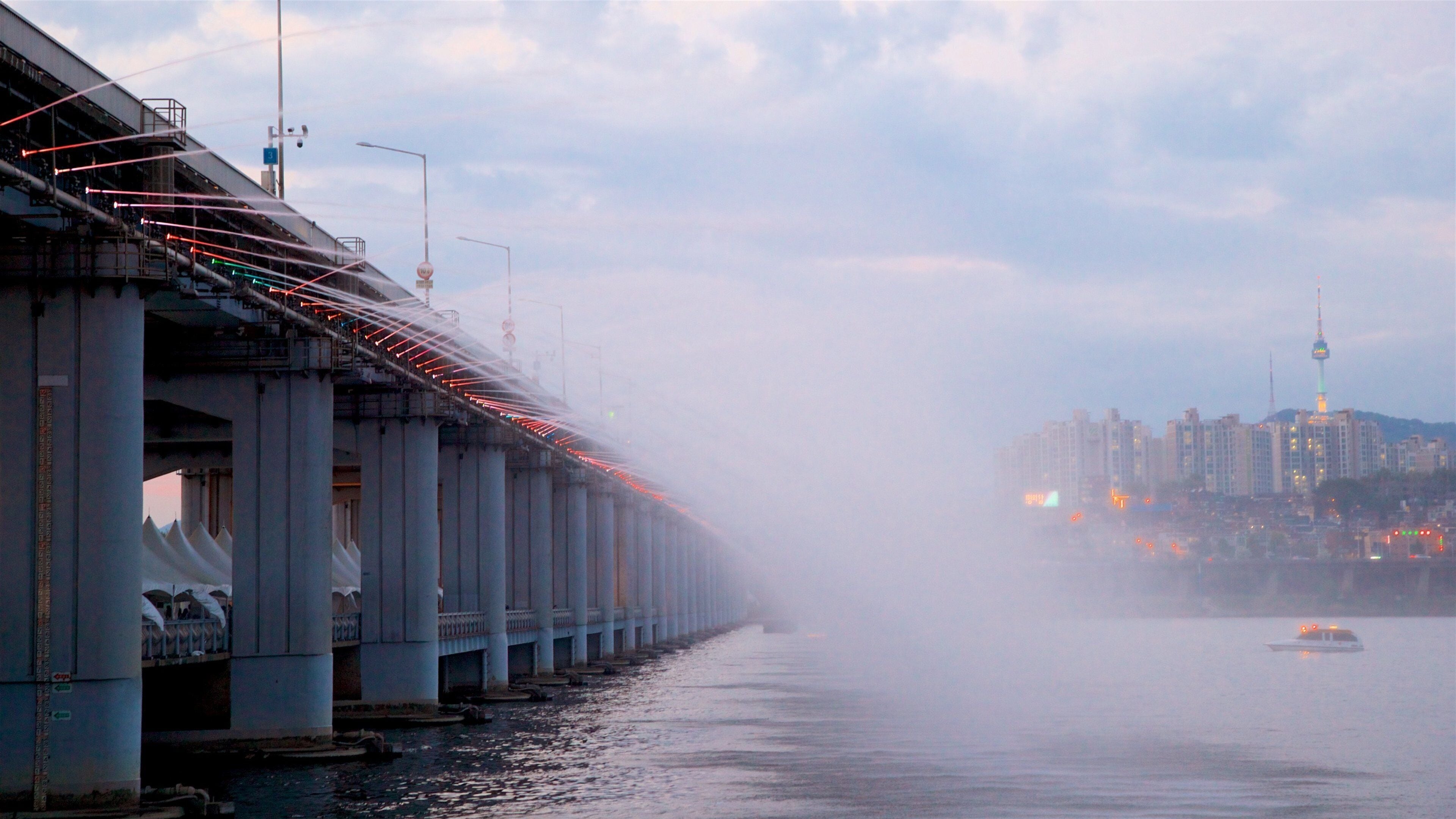 A bridge with red lights, fog, and a cityscape with buildings and a tower in the background.