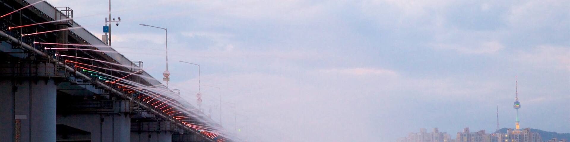 A bridge with red lights, fog, and a cityscape with buildings and a tower in the background.