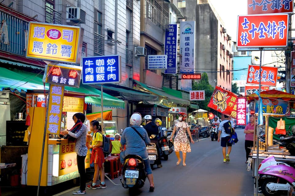 Un marché de rue animé, avec de nombreux étals de nourriture et des panneaux en langue étrangère.