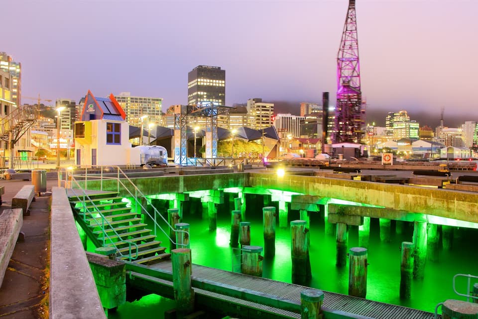 A waterfront with illuminated green water, a dock, and a cityscape with buildings and a crane in the background.