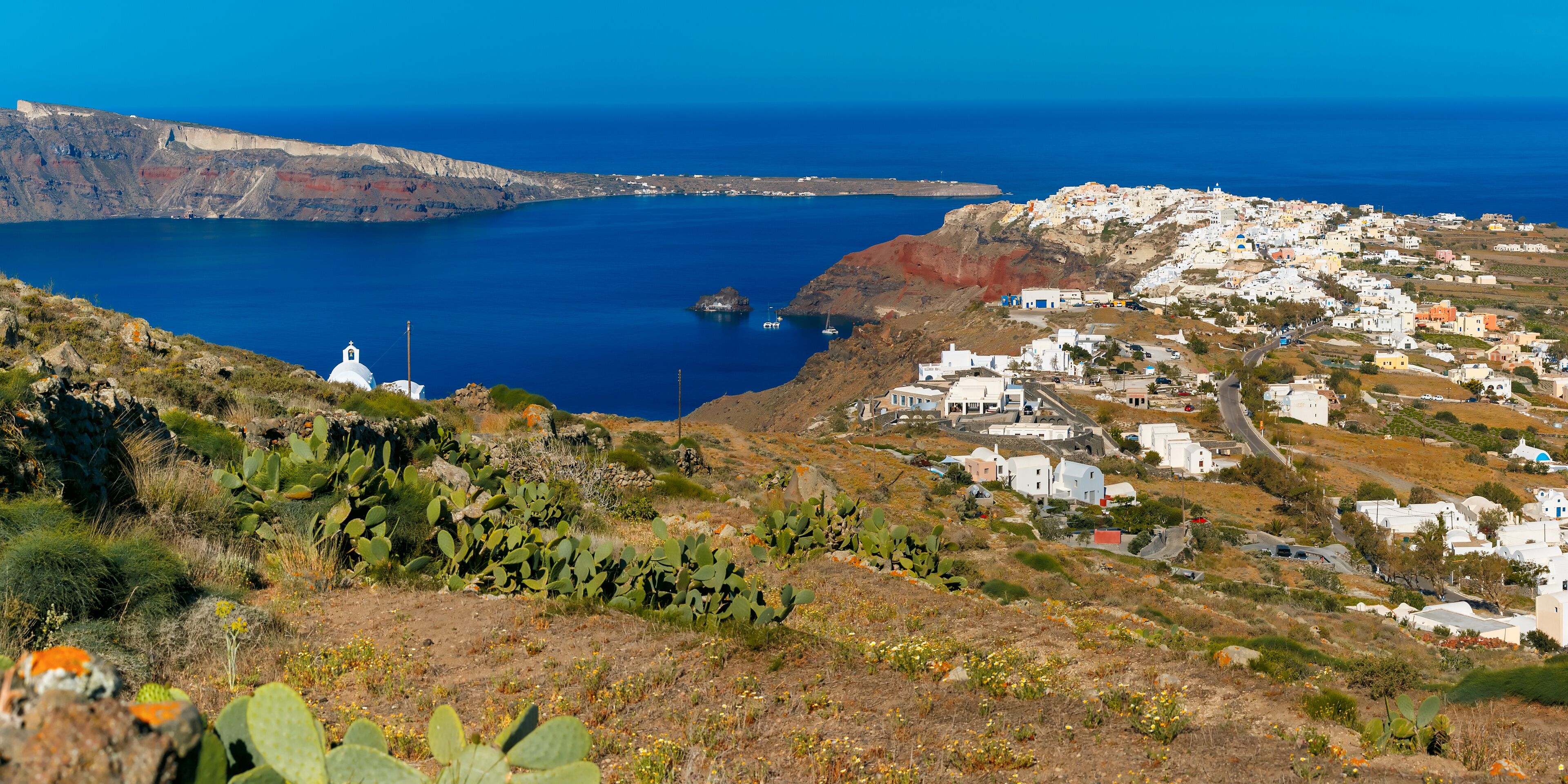A coastal town with white buildings, a clear blue sea, and a rocky landscape with cacti.