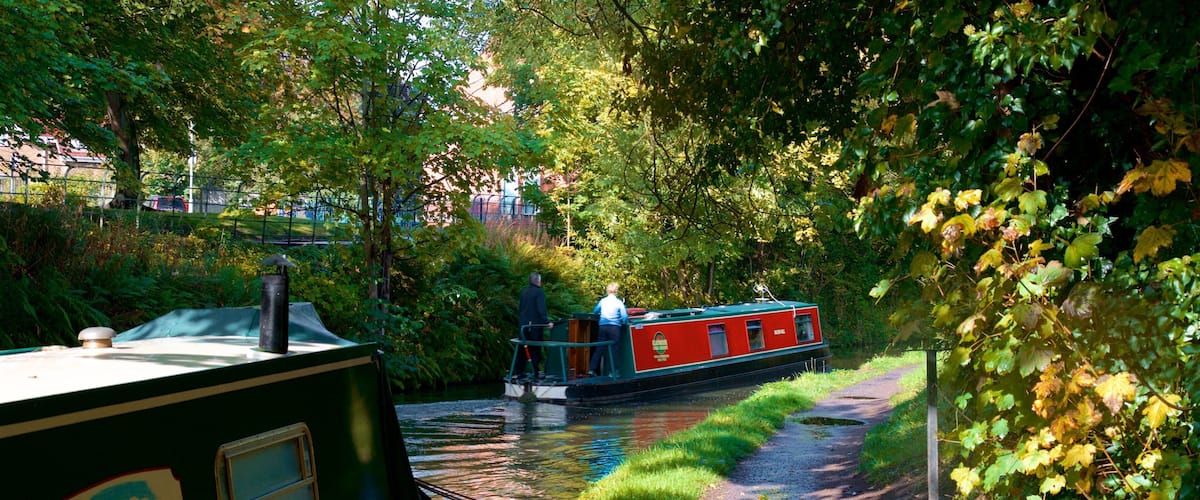 A narrow waterway with a narrowboat named 'Moorehen' docked on the left, a red narrowboat on the right, and a path alongside the water.